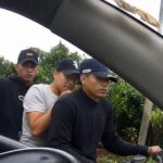 Hombre acompañado por agentes durante un procedimiento junto a un vehículo.