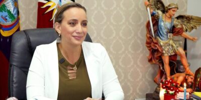 Johana Núñez sentada en una oficina con una bandera ecuatoriana al fondo.