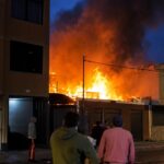 Llamas y humo junto a una vivienda al anochecer, con personas en la calle.