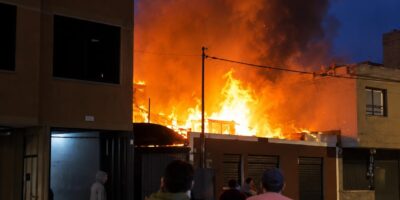 Llamas y humo junto a una vivienda al anochecer, con personas en la calle.