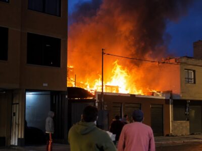 Llamas y humo junto a una vivienda al anochecer, con personas en la calle.