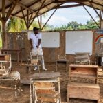 Estudiantes y pupitres en un aula rural con techo de madera y paredes abiertas.