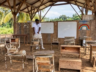 Estudiantes y pupitres en un aula rural con techo de madera y paredes abiertas.