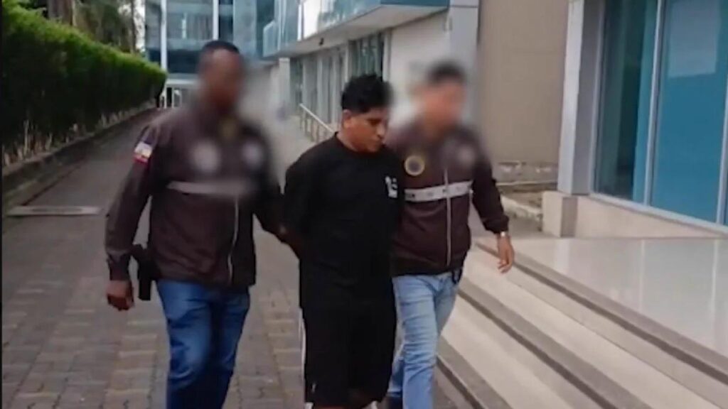 Dos agentes acompañan a un hombre frente a un edificio; los rostros de los agentes están difuminados.