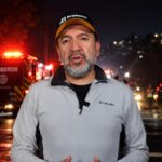 Un integrante del Cuerpo de Bomberos habla ante la cámara con unidades de emergencia al fondo.