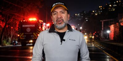 Un integrante del Cuerpo de Bomberos habla ante la cámara con unidades de emergencia al fondo.