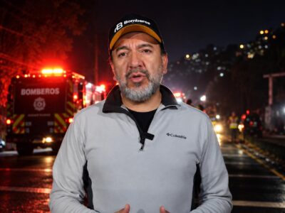 Un integrante del Cuerpo de Bomberos habla ante la cámara con unidades de emergencia al fondo.