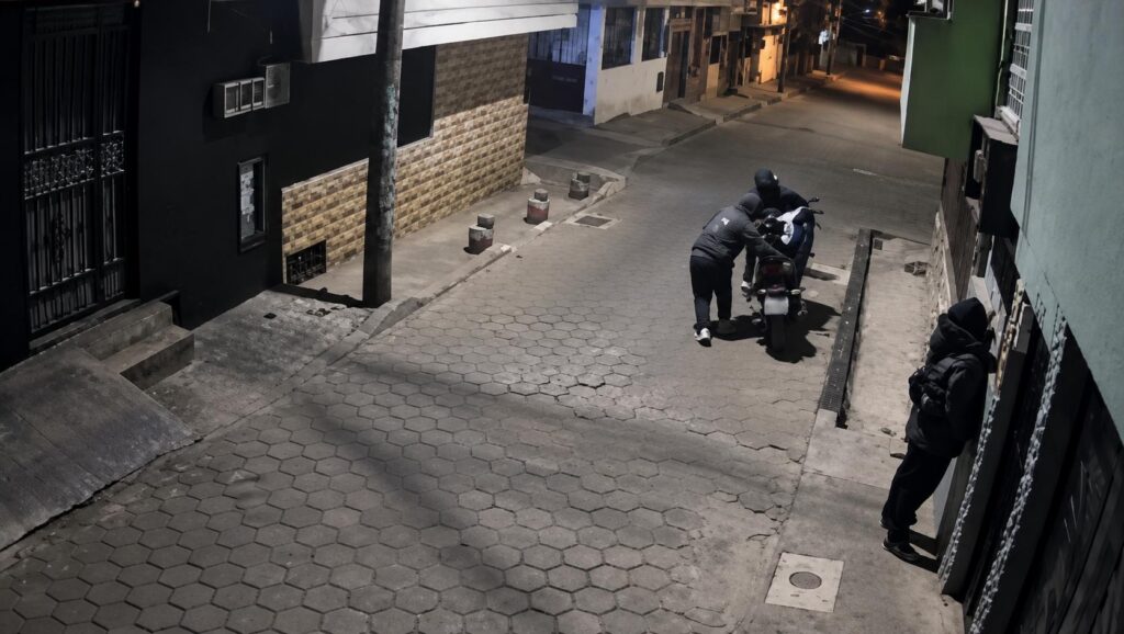 Captura de una cámara de seguridad con personas junto a una motocicleta en una acera durante la noche.