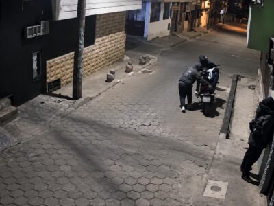 Captura de una cámara de seguridad con personas junto a una motocicleta en una acera durante la noche.