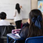 Estudiantes sentados en pupitres frente a una docente en un aula.