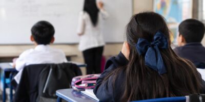 Estudiantes sentados en pupitres frente a una docente en un aula.