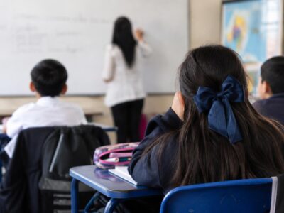 Estudiantes sentados en pupitres frente a una docente en un aula.