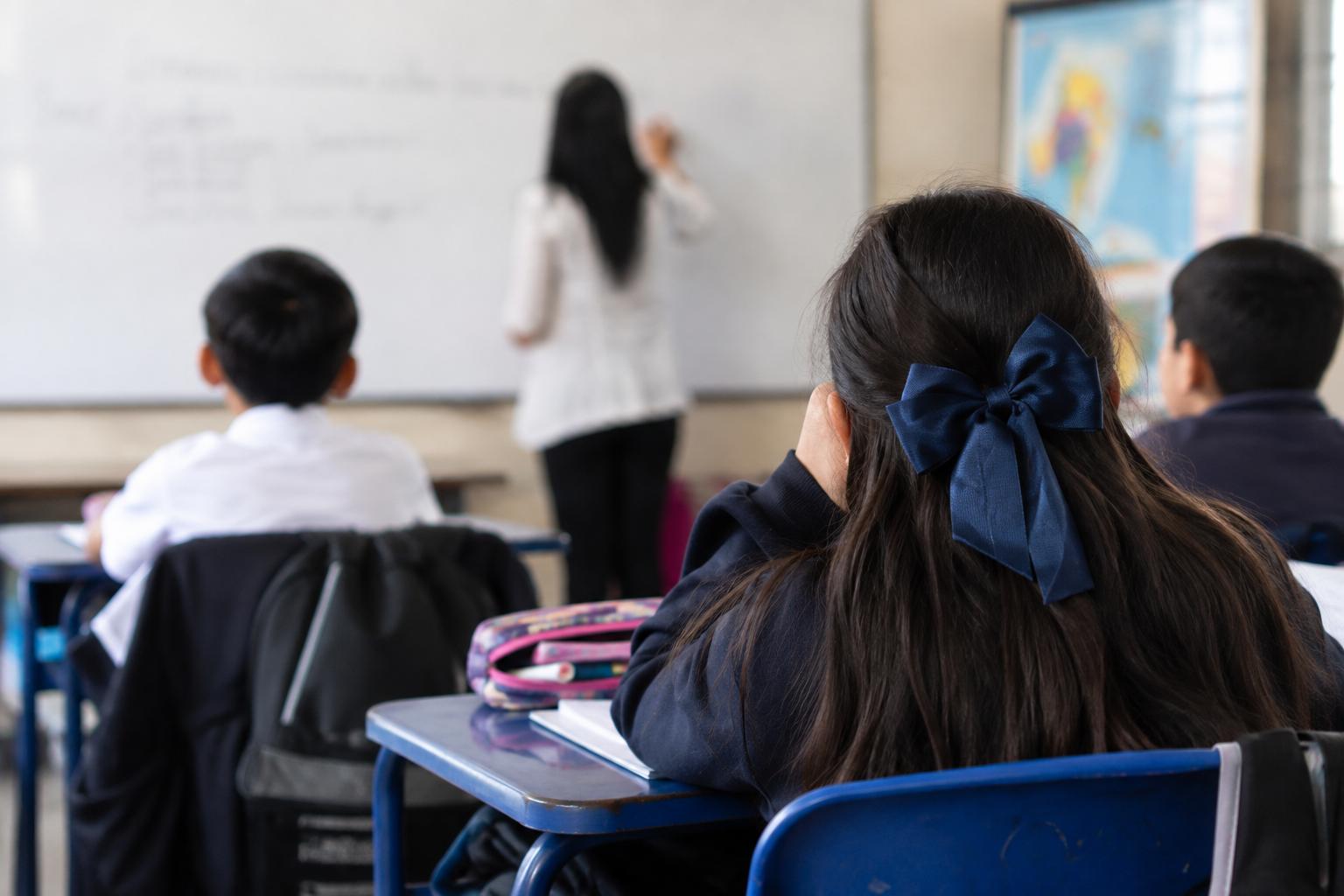 Estudiantes sentados en pupitres frente a una docente en un aula.