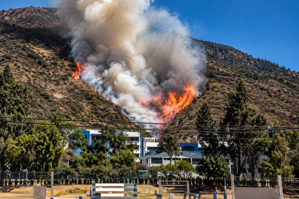 Humo y llamas en una ladera, con viviendas y árboles en primer plano.