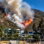 Humo y llamas en una ladera, con viviendas y árboles en primer plano.