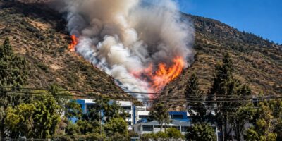 Humo y llamas en una ladera, con viviendas y árboles en primer plano.