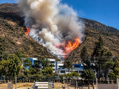 Humo y llamas en una ladera, con viviendas y árboles en primer plano.