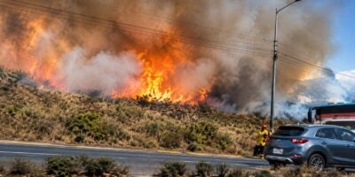 Llamas y humo sobre una ladera junto a una vía con vehículos.