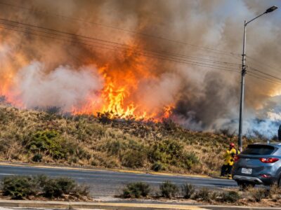 Llamas y humo sobre una ladera junto a una vía con vehículos.