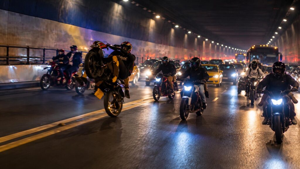 Motociclistas y otros vehículos dentro de un túnel iluminado durante la noche.