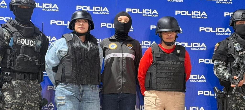 Agentes de Policía flanquean a dos mujeres con chalecos y cascos frente a un panel institucional.