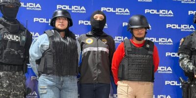 Agentes de Policía flanquean a dos mujeres con chalecos y cascos frente a un panel institucional.