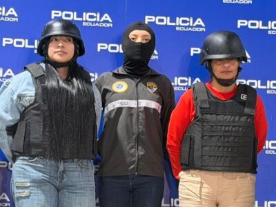 Agentes de Policía flanquean a dos mujeres con chalecos y cascos frente a un panel institucional.
