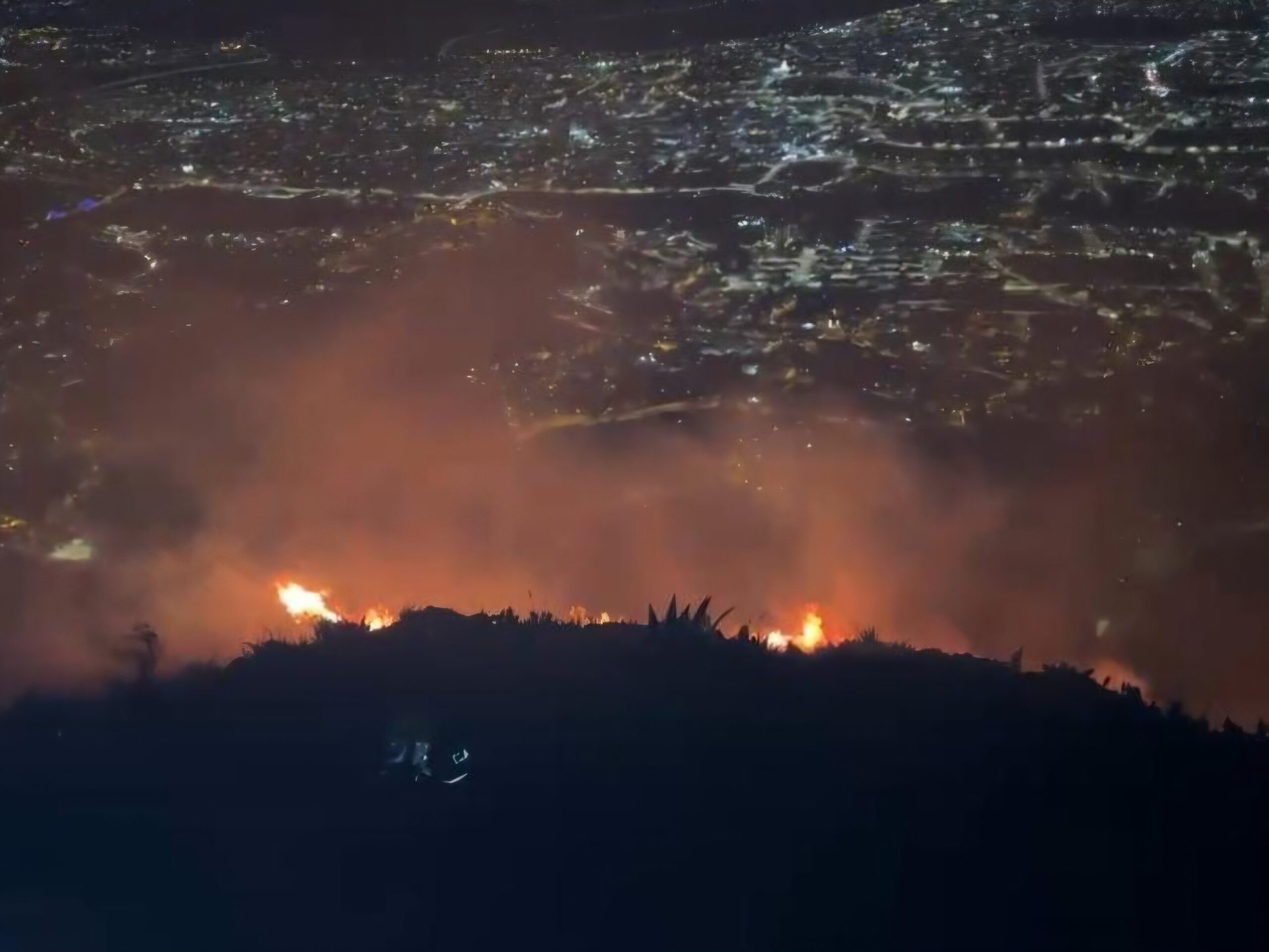 Llamas del incendio forestal en el caserío Apagua, parroquia Ambatillo, con la ciudad de Ambato iluminada al fondo, la noche del 6 de octubre de 2026.