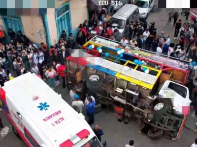 Un vehículo de transporte volcado en una calle, rodeado de personas y una ambulancia.