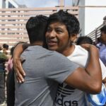 Dos hombres se abrazan en un espacio público, con otras personas al fondo.