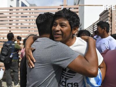 Dos hombres se abrazan en un espacio público, con otras personas al fondo.