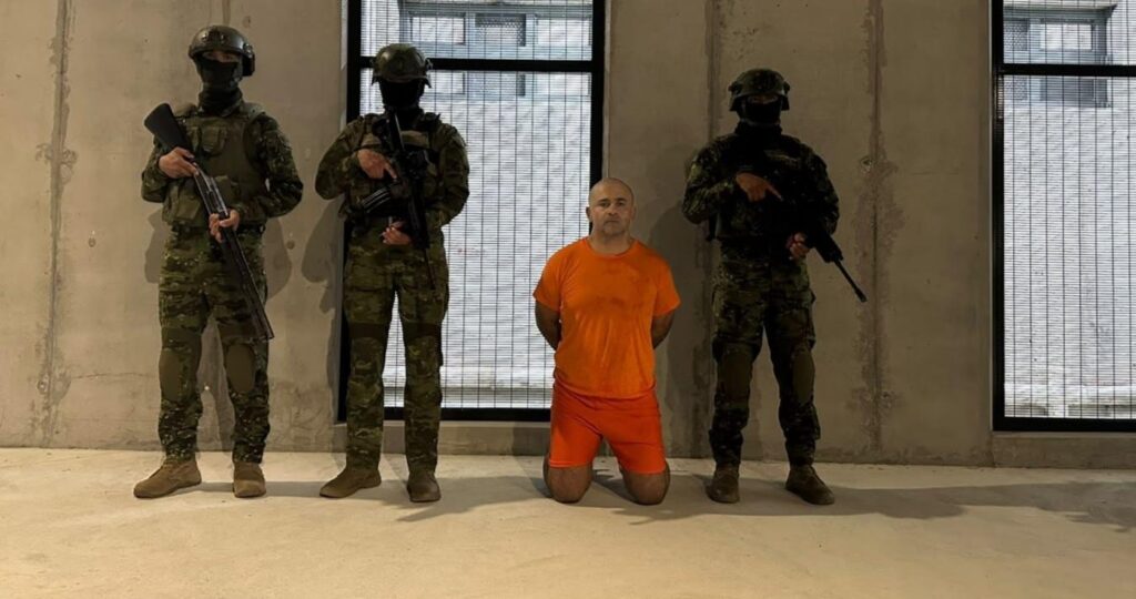 Un hombre con uniforme naranja arrodillado entre agentes armados en el interior de un edificio.