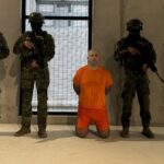 Un hombre con uniforme naranja arrodillado entre agentes armados en el interior de un edificio.