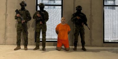 Un hombre con uniforme naranja arrodillado entre agentes armados en el interior de un edificio.