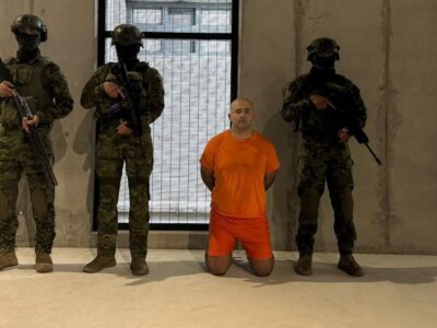Un hombre con uniforme naranja arrodillado entre agentes armados en el interior de un edificio.