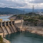 Imagen ilustrativa de una represa con bajo nivel de agua, torres eléctricas y un complejo industrial al fondo.