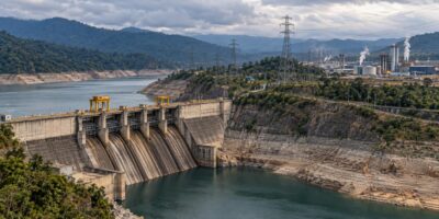 Imagen ilustrativa de una represa con bajo nivel de agua, torres eléctricas y un complejo industrial al fondo.